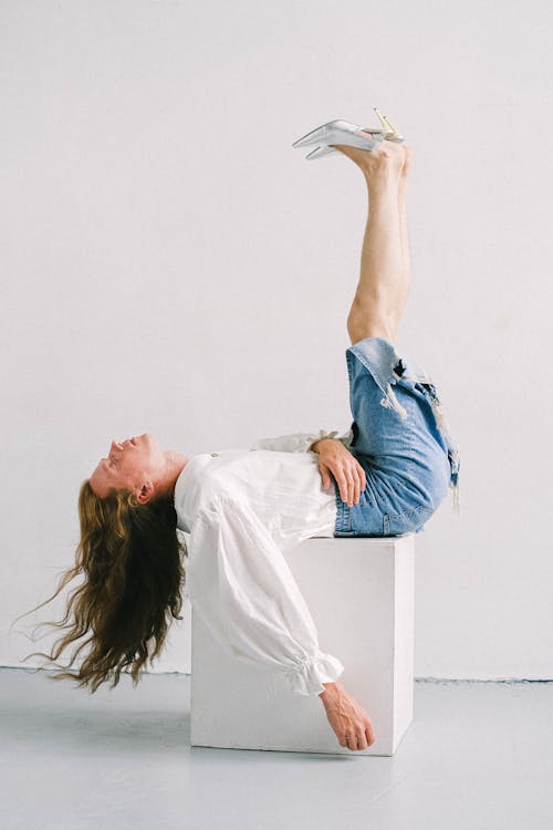 Free Side view of young eccentric androgynous man with long wavy hair in feminine outfit lying on cube with raised legs in white studio Stock Photo