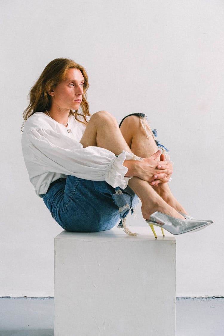 Serious Androgynous Man Embracing Knees On Cube In Studio