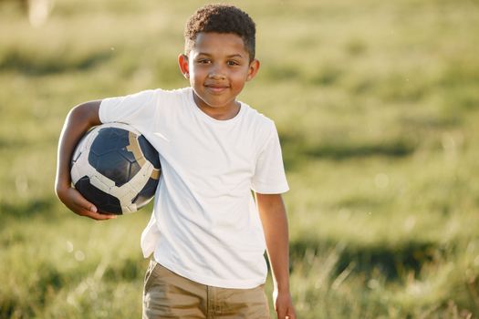 Young boy in a white shirt smiling while holding a soccer ball outdoors.