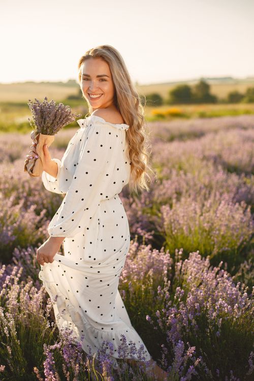 Free Caucasian woman in a polka dot dress smiling while holding a lavender bouquet in a scenic field at sunset. Stock Photo