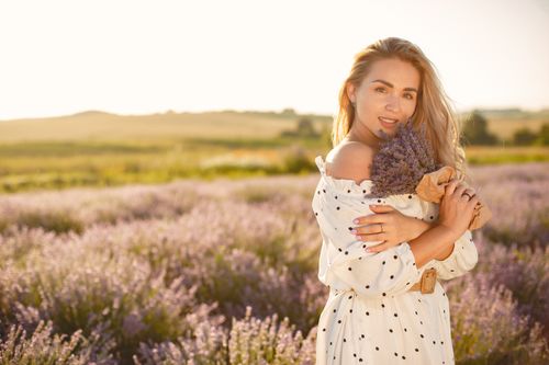 Free Blonde woman in a white dress poses with a lavender bouquet in a sunlit field. Perfect for summertime stock photos. Stock Photo