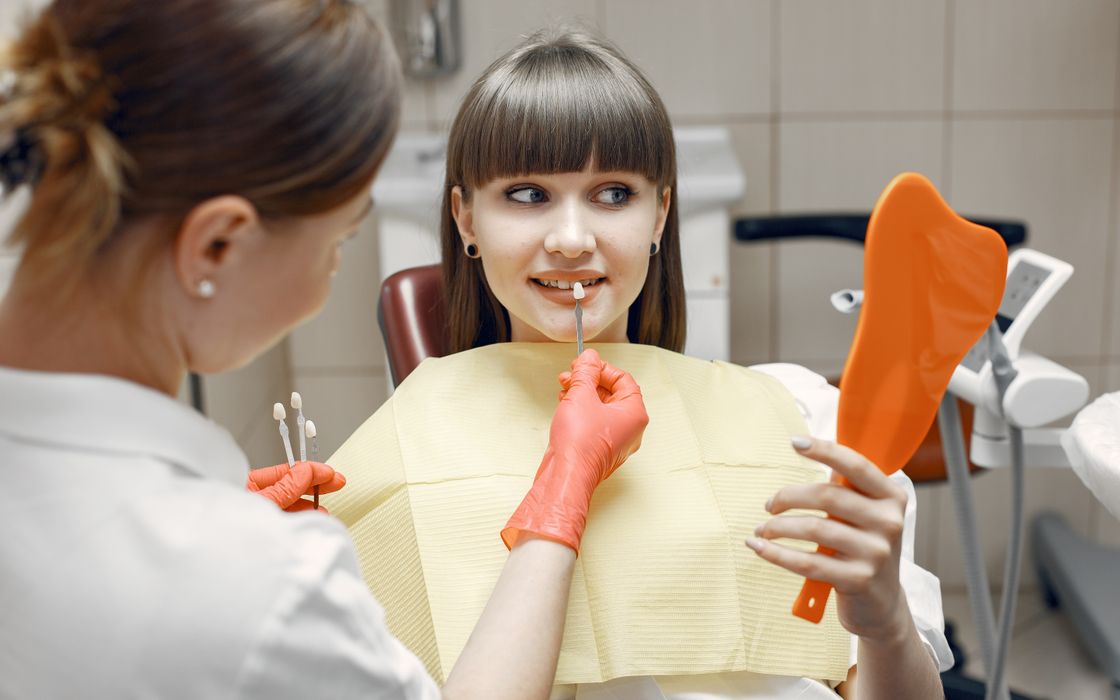 Dentist examining a young woman's front teeth while she holds a hand mirror during a consultation