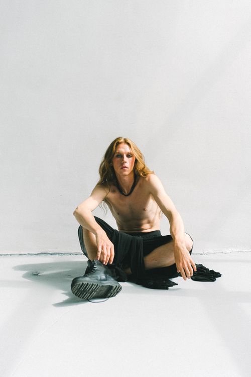 Free Full body of young slim shirtless gender fluid male with long wavy hair in black skirt and boots sitting on floor and looking at camera against white background Stock Photo