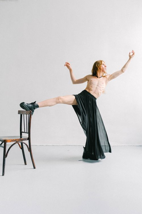 Free Full body of young concentrated slender shirtless androgynous male with reddish hair in long black skirt raising arms and stretching leg in bright room in daylight Stock Photo