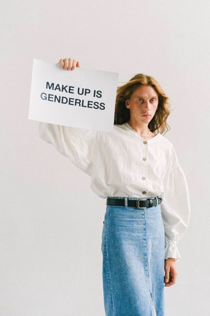 Thoughtful young man with reddish hair in white blouse and denim skirt looking at camera while demonstrating sheet with inscription Make Up Is Genderless on white background