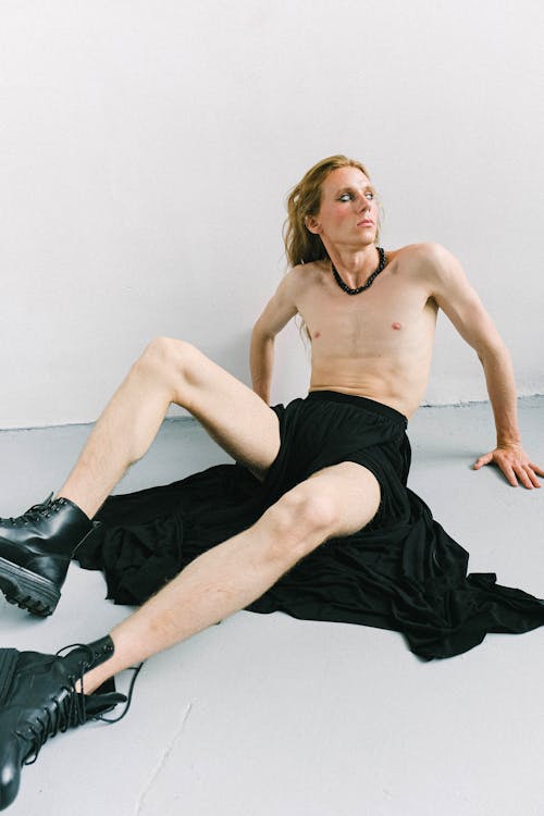 Full body of dreamy young handsome shirtless man in long black skirt and boots leaning on floor and thoughtfully looking away while lounging in bright room in daylight