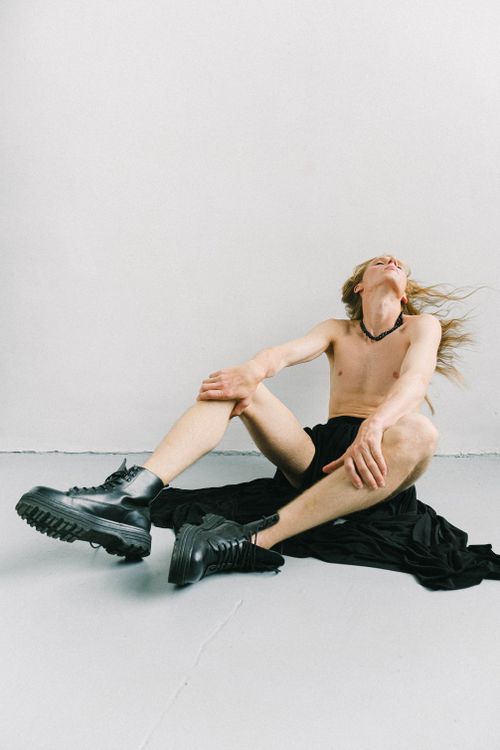 Free Full body of pensive young slim shirtless male with long flying hair in long skirt and boots looking up and touching legs while resting in bright studio in daylight Stock Photo