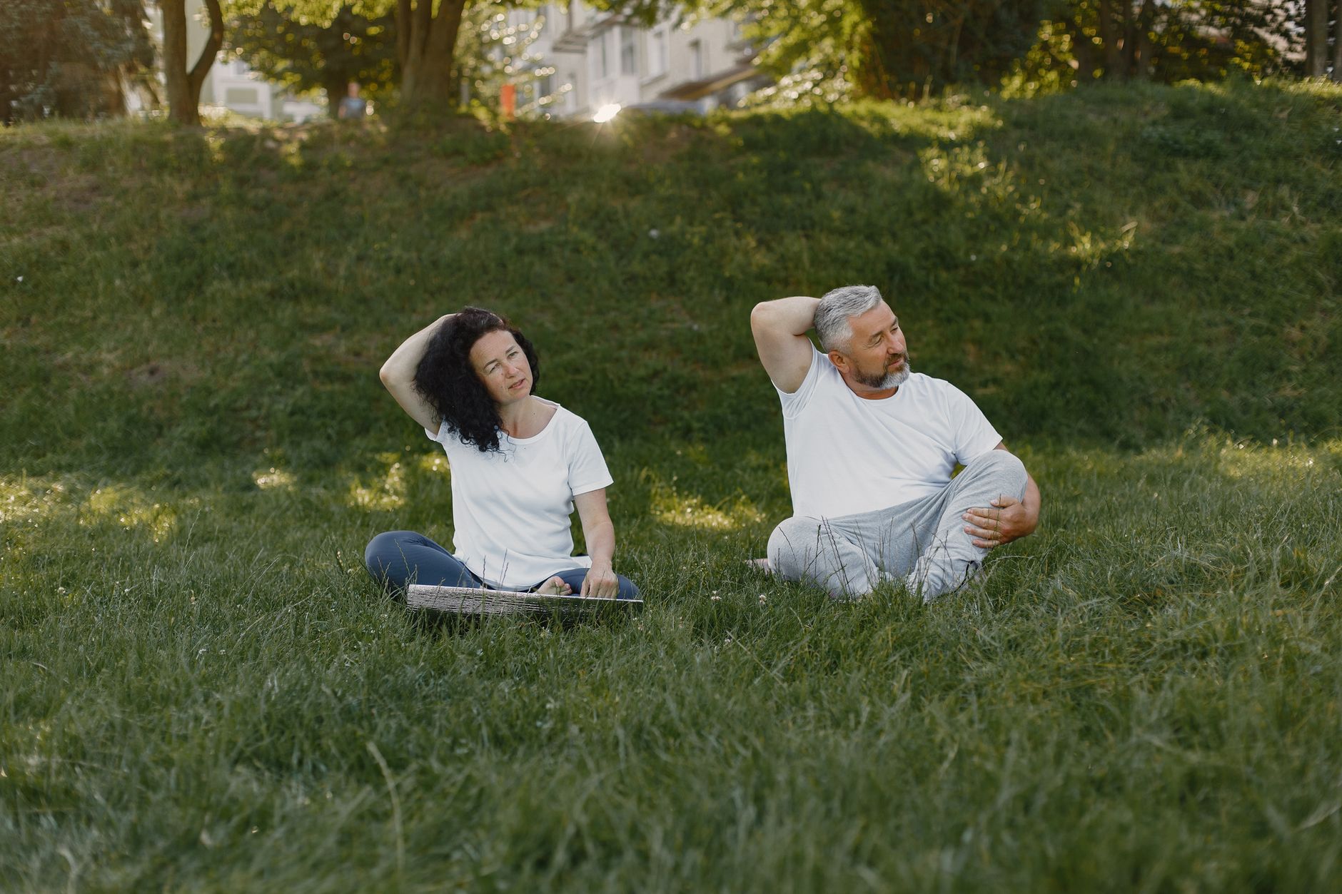 Couple doing neck stretches outdoors, promoting a healthy lifestyle in a sunny park setting.