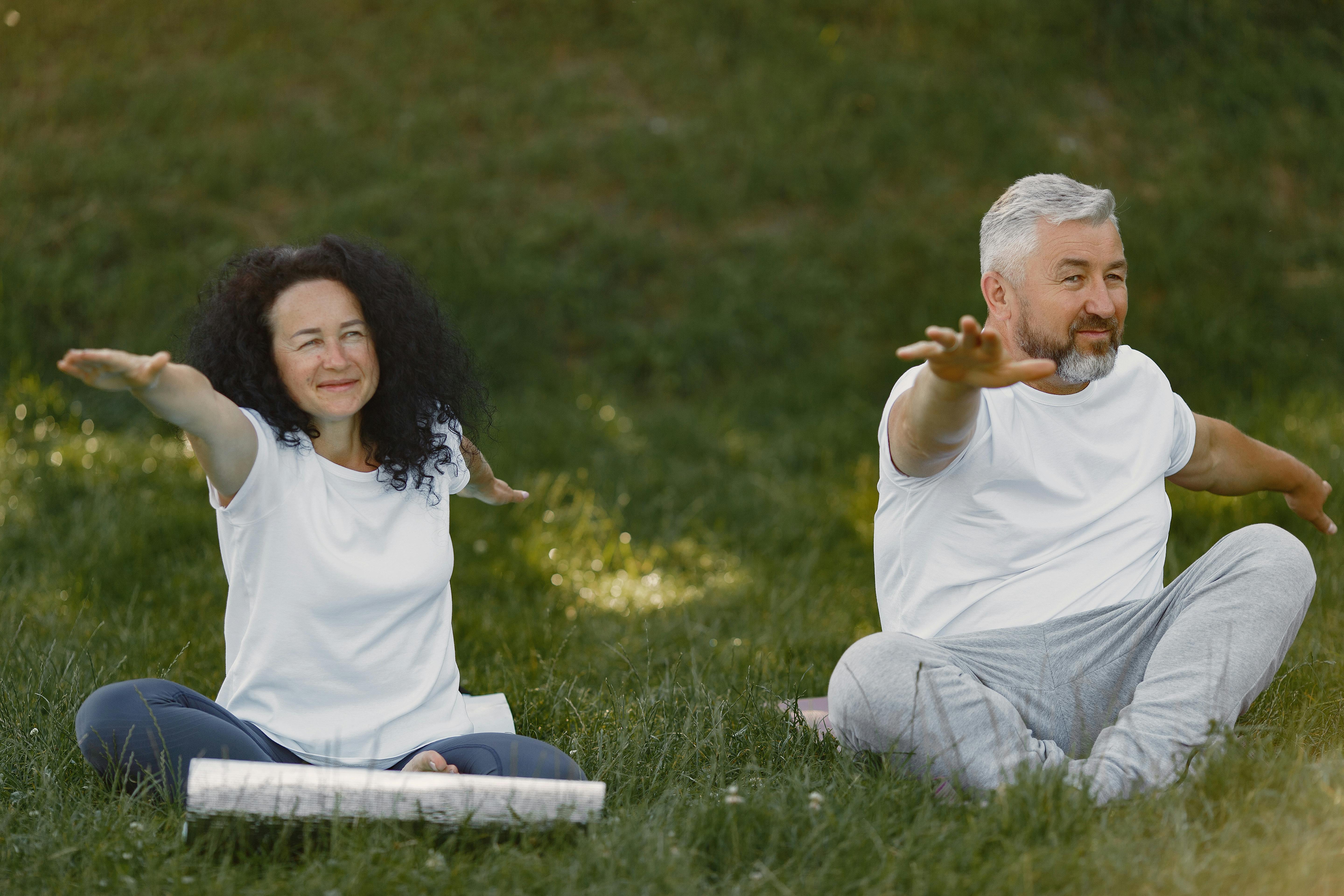 Elderly couple doing yoga exercises on grass in a park, promoting wellness and fitness.