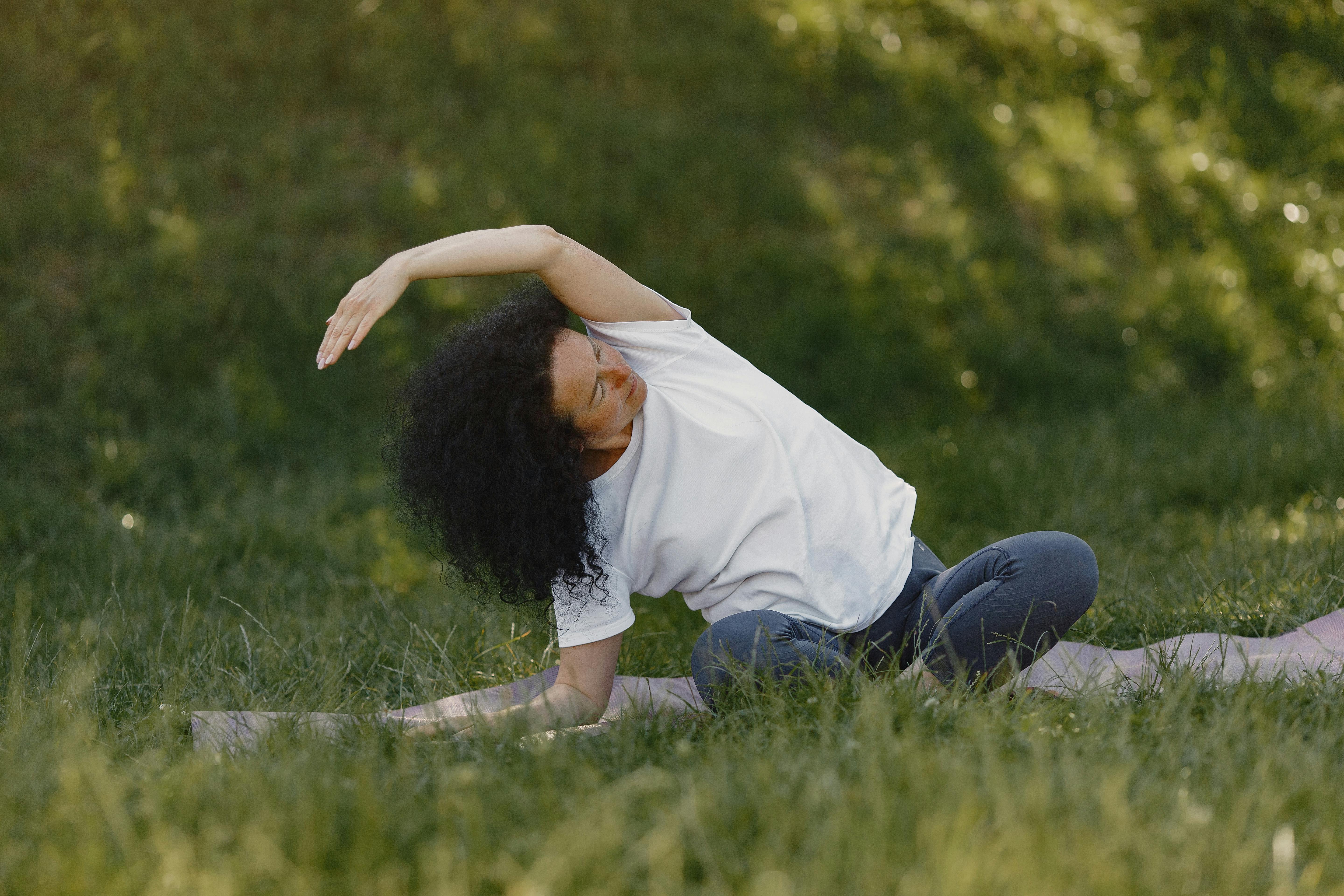 A woman in a white shirt practicing yoga outdoors on green grass, showcasing stretching and wellness.