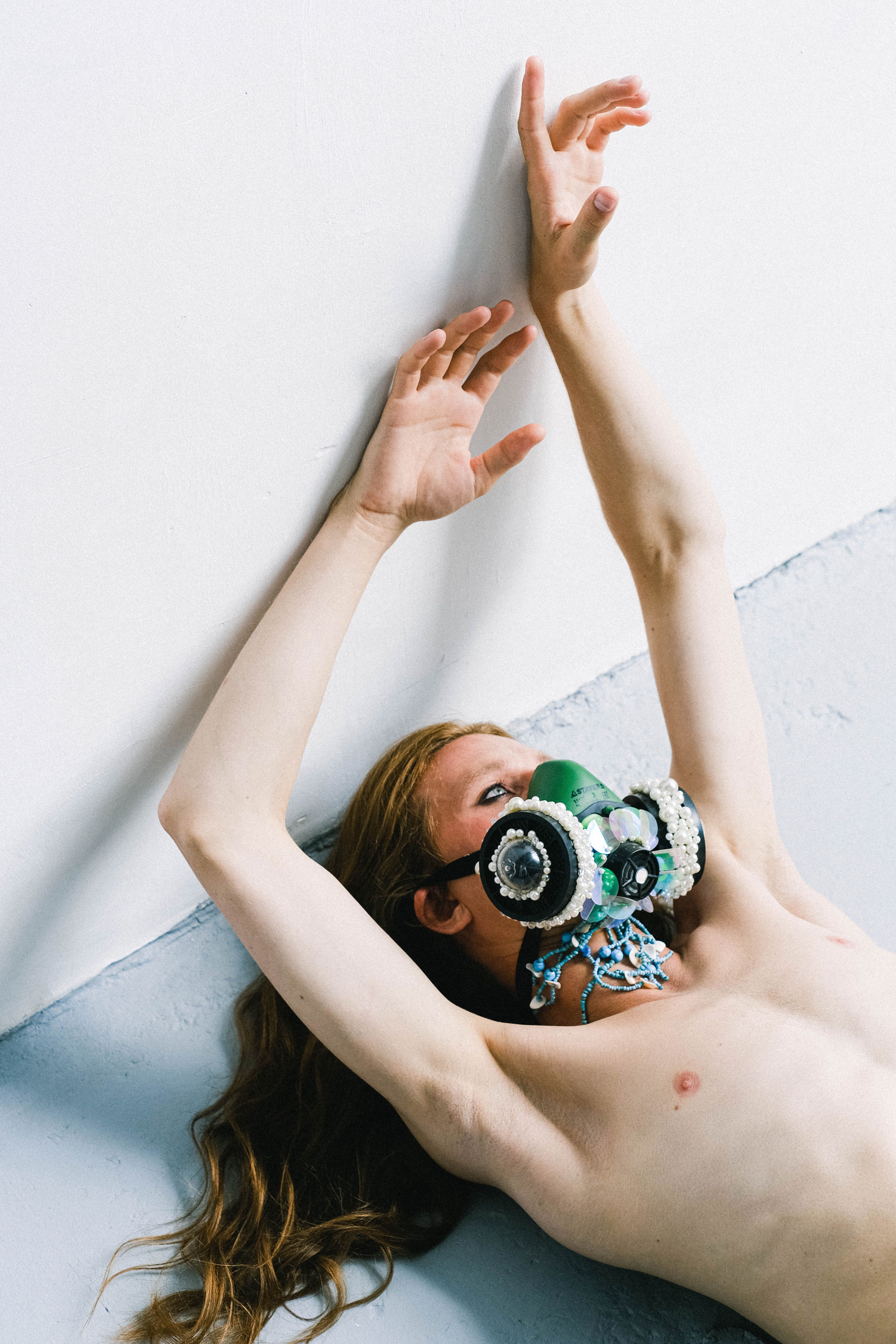 From above glam shirtless male with long ginger hair wearing green unusual respirator decorated with pearls while lying on floor with raised skinny arms and looking away