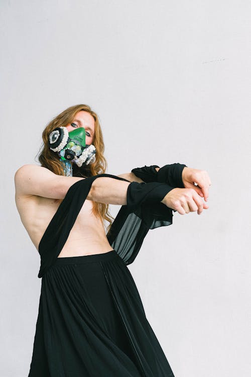 Androgynous shirtless male model with long ginger hair wearing black skirt and creative respirator decorated with white pearls standing with arms outstretched against white wall and looking at camera