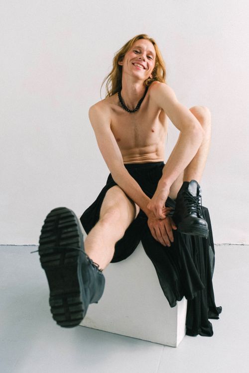 Free Full body Smiling shirtless male model with long hair wearing black maxi skirt and massive boots sitting on cube in light studio Stock Photo