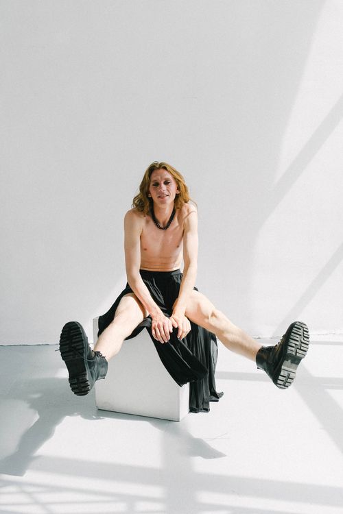 Free Full length positive androgynous male with naked torso and long ginger hair wearing black maxi skirt and massive boots while sitting on cube with legs raised and looking at camera Stock Photo