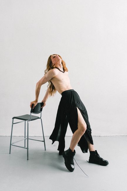 Shirtless model in a skirt and boots striking a pose in a studio setting.