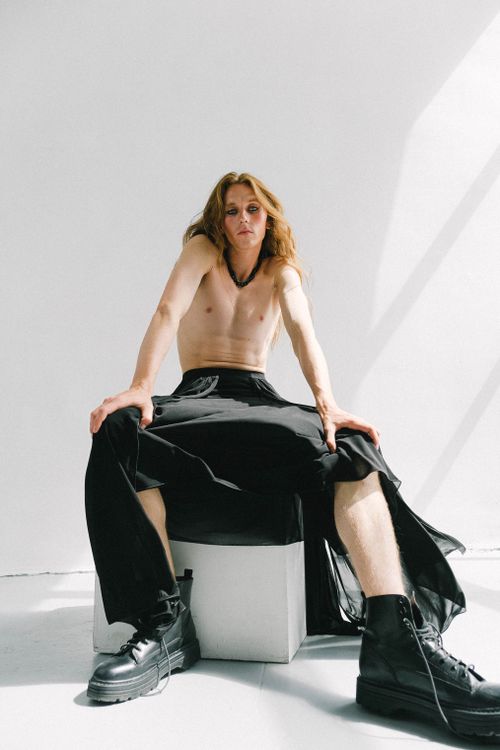 Free Eccentric androgynous male model with naked torso and long ginger hair wearing black maxi skirt and massive boots sitting on cube in light studio Stock Photo