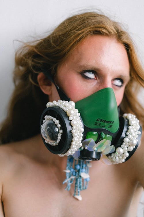 Anonymous eccentric shirtless androgynous man with long hair in stylish respirator covered with pearls rolling eyes against white background