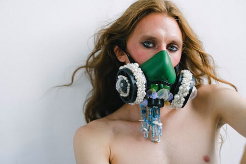 Unrecognizable confident young androgynous man with naked body and long wavy hair wearing fashionable respirator mask decorated with beads standing against white background and looking at camera