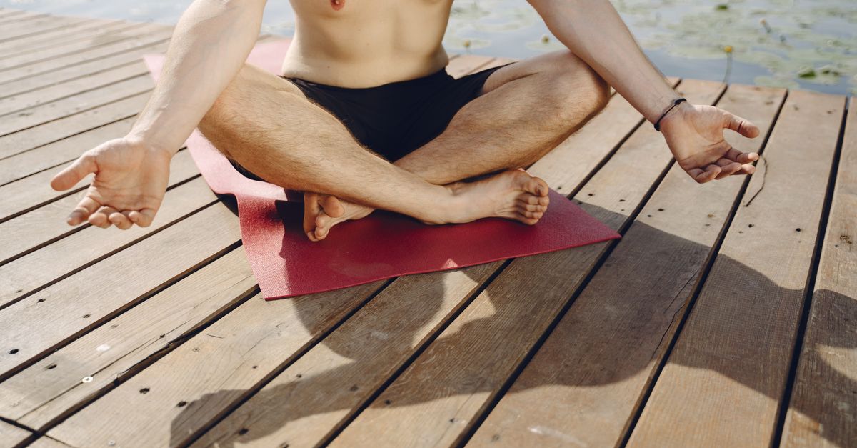 A man practices meditation on a wooden pier by the water, seated on a red yoga mat under sunlight.