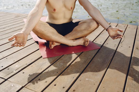 A man practices meditation on a wooden pier by the water, seated on a red yoga mat under sunlight.