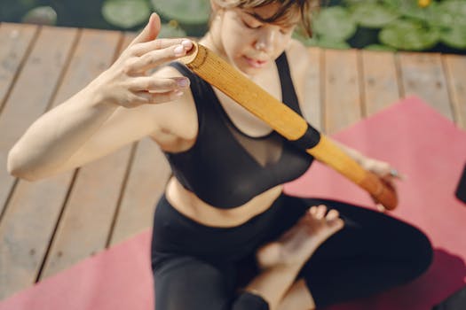 Woman meditating outdoors on yoga mat in serene environment using a wooden stick.