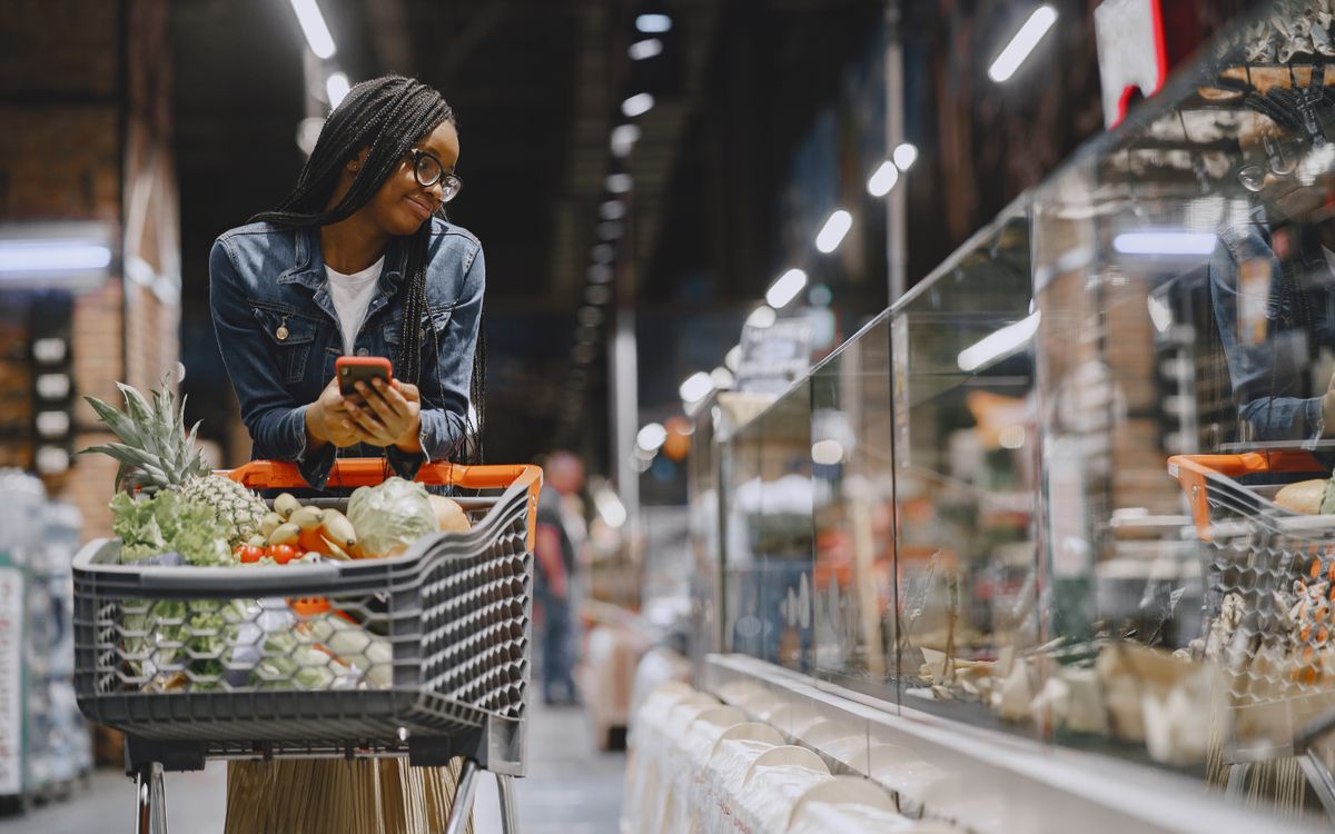 A young woman with afro braids grocery shopping in a supermarket, using a mobile phone.
