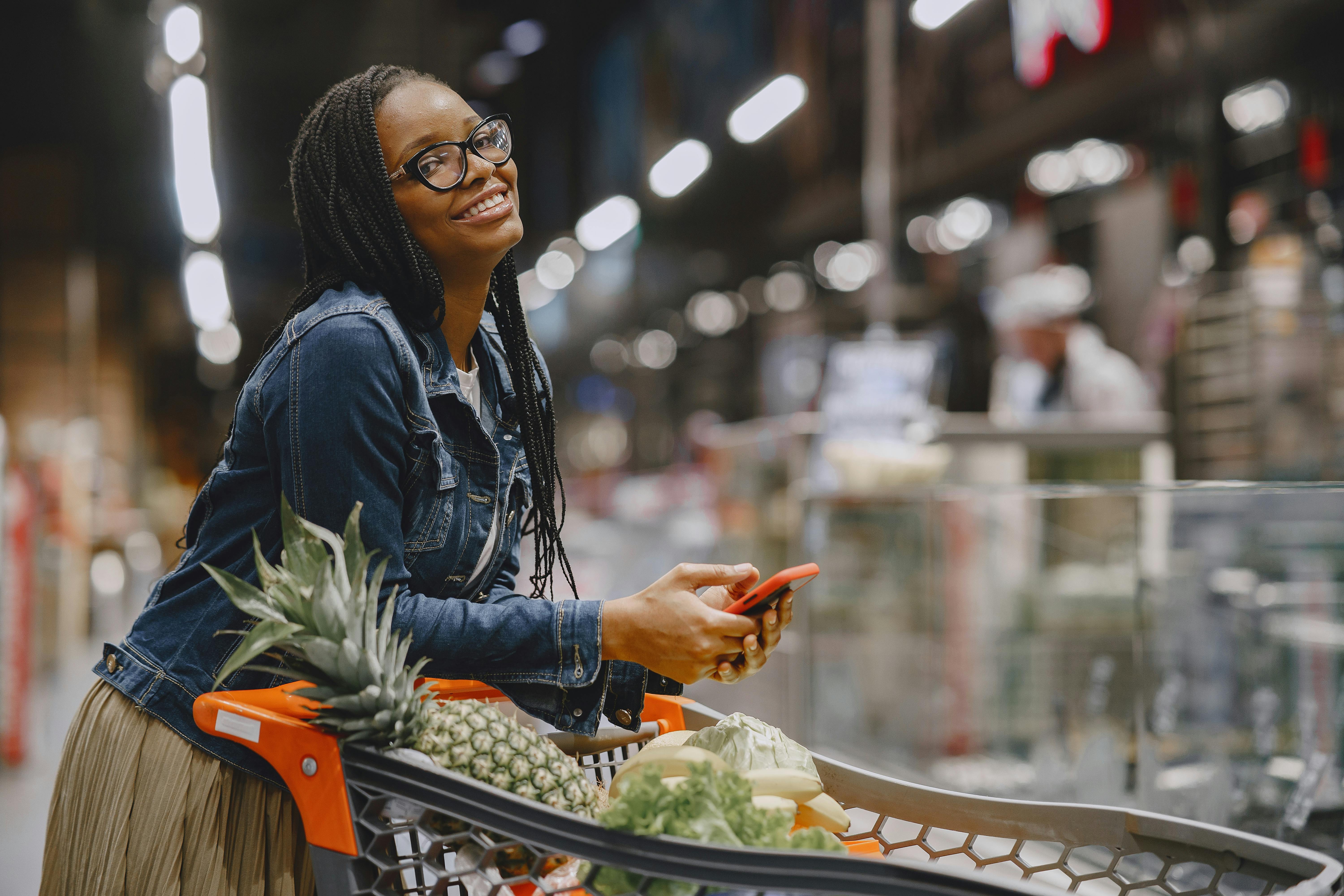 Smiling woman with smartphone shopping in supermarket aisle, using cart.