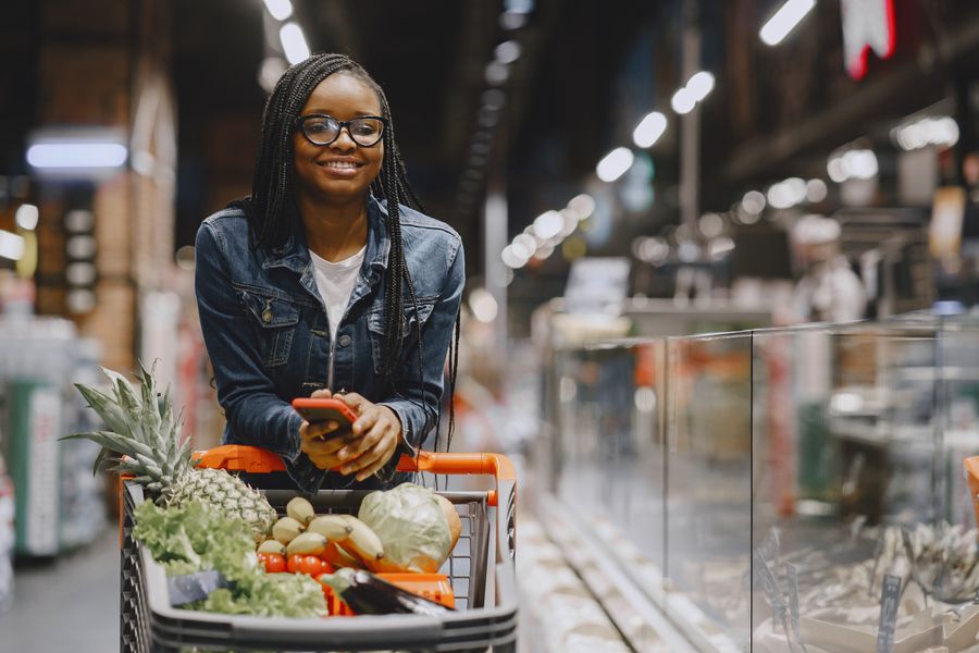 A woman smiles while shopping for fresh produce in a supermarket, using a smartphone.