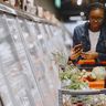 A young woman looks seriously at her smartphone while pushing a shopping cart down a supermarket aisle.
