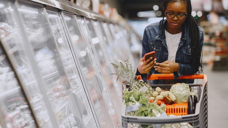 Woman in a grocery store using smartphone while pushing a shopping cart filled with groceries.