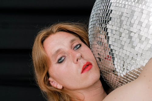 Pensive young androgynous man with long hair and makeup hugging mirrored disco ball and looking at camera against black background