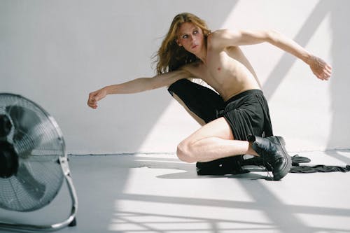 Eccentric shirtless young androgynous man sitting sitting on haunches near fan in studio