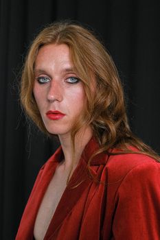 一位神情严肃的年轻男女模特化着妆，留着姜黄色长发，身穿红色西装外套，站在黑色背景前，若有所思地望向镜头
