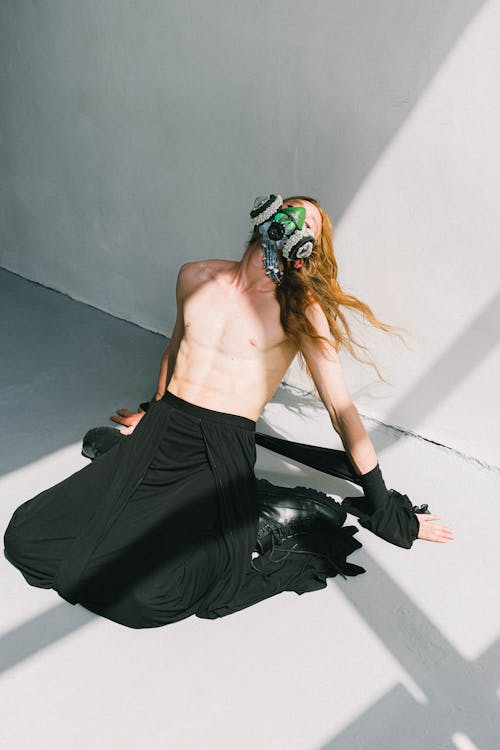 From above of unrecognizable fit shirtless androgynous man with long ginger hair in maxi skirt and creative respirator mask sitting on floor with closed eyes