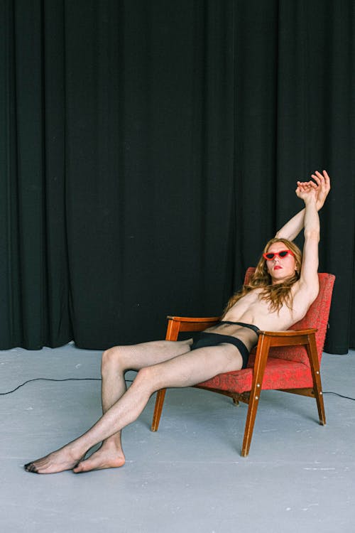 Full body of sensual self assured androgynous male with long hair and naked torso in panties and tights sitting in armchair with hands over head against black curtain