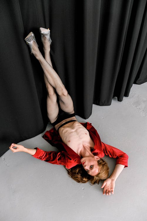 From above of extravagant skinny young transsexual male dancer in provocative clothes and high heeled shoes lying on floor with raised legs near black curtain