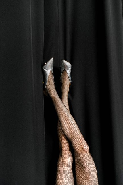 Legs of crop unrecognizable dancer in black fishnet tights and high heeled shoes against black curtain