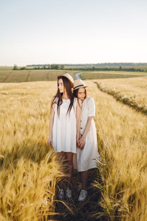 Two Women in White Dress and Sun Hat Holding Hands in a Green Grass Field