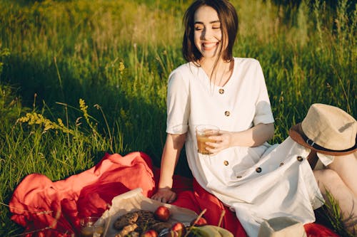 Woman in White Dress Holding a Glass of Drink on a Red Picnic Blanket