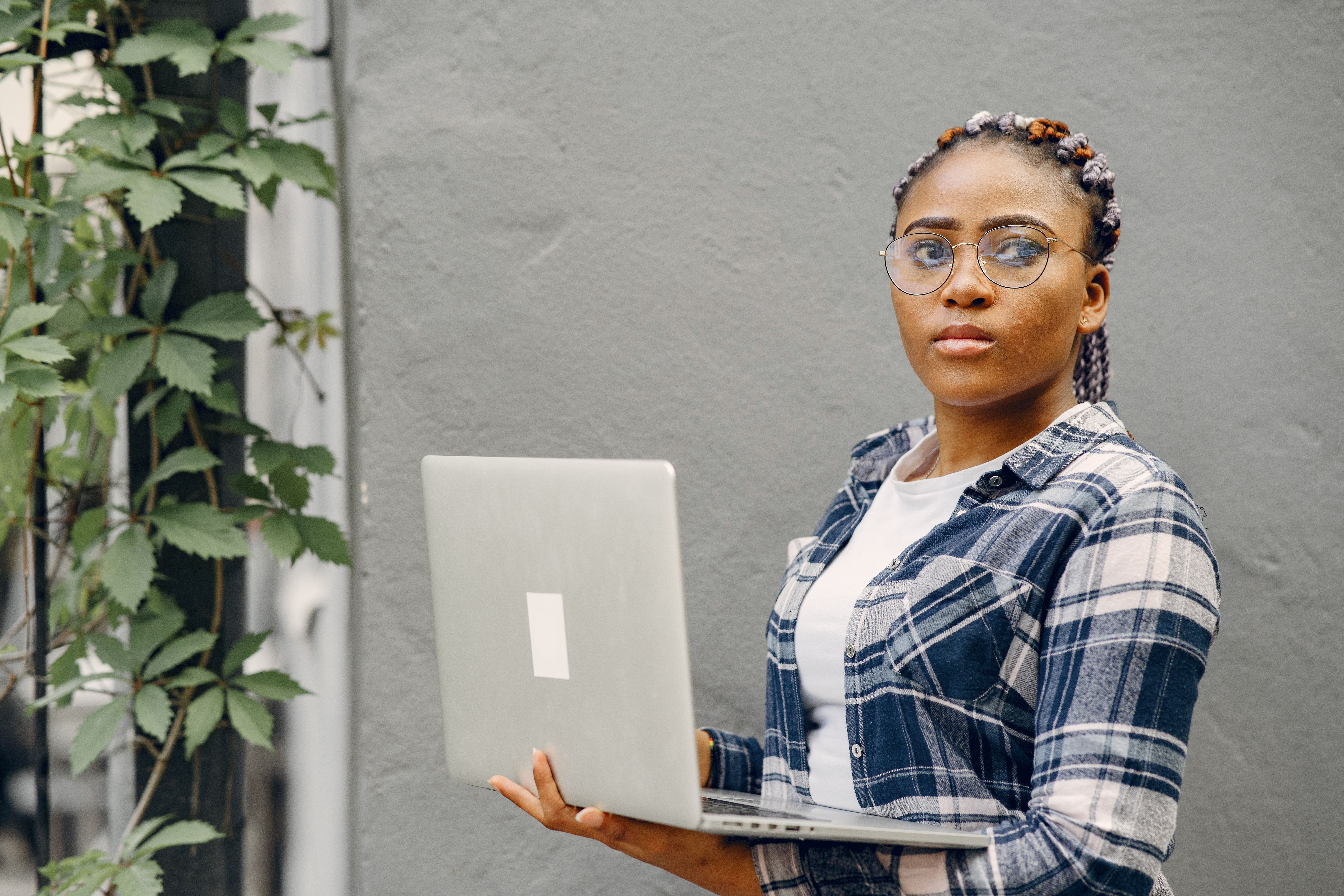 Woman wearing glasses holds a laptop indoors beside a concrete wall, checking a wireless connection.