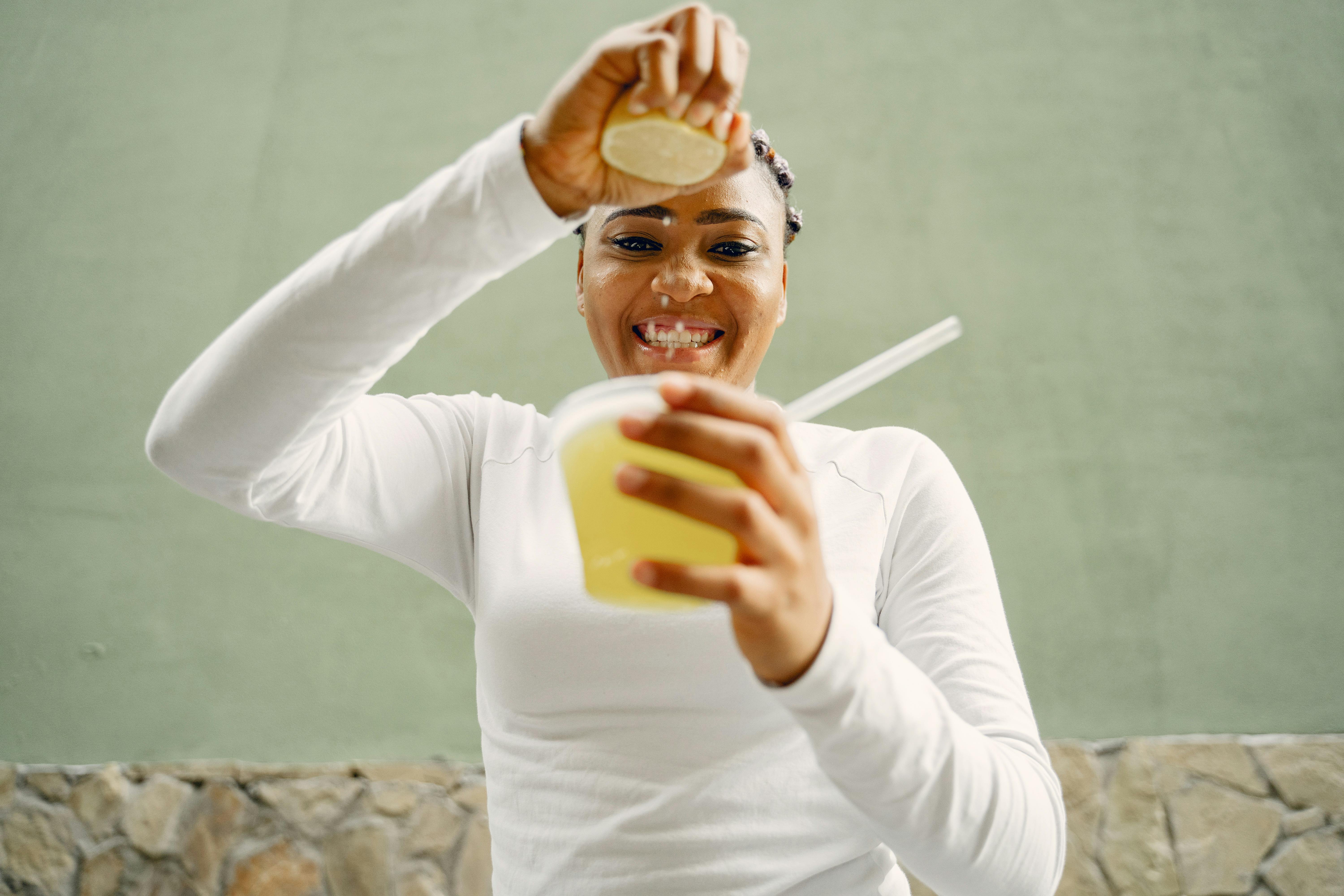 Happy woman in white squeezing fresh lemon into lemonade cup outdoors.