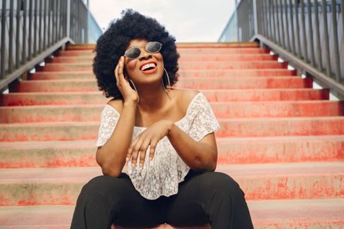 Free Joyful woman wearing sunglasses and afro hairstyle sitting outdoors on red steps. Stock Photo