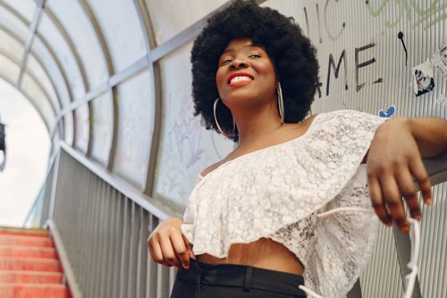 Free Confident woman in a crop top with an afro hairstyle smiles brightly while leaning on a stairway railing. Stock Photo