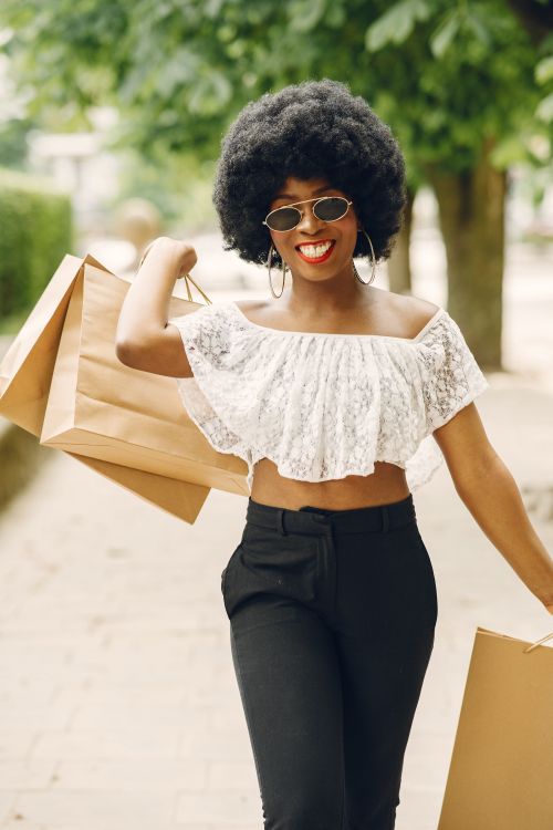 Free A fashionable woman with afro hair smiling and carrying shopping bags on a sunny day. Stock Photo