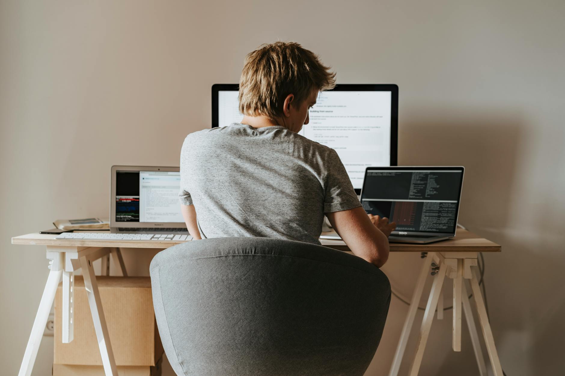 Young man coding at home office with multiple laptops, representing remote work and technology.