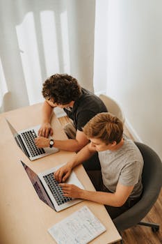 Two men collaborating on laptops at a table, focused on programming tasks.