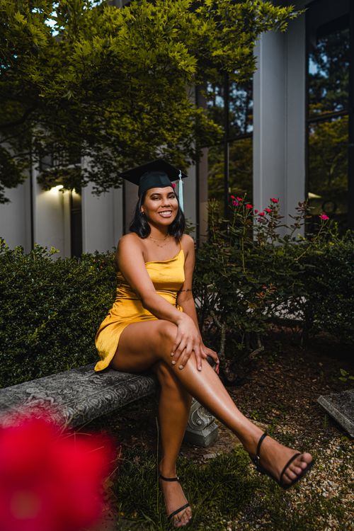 Free Full body of optimistic female in graduation cap and yellow dress sitting with legs crossed on stone parapet near green bushes with flowers and looking at camera Stock Photo