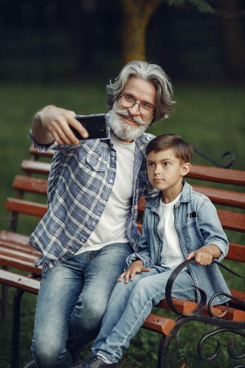 Free A senior man and young boy smiling and taking a selfie on a park bench. Stock Photo