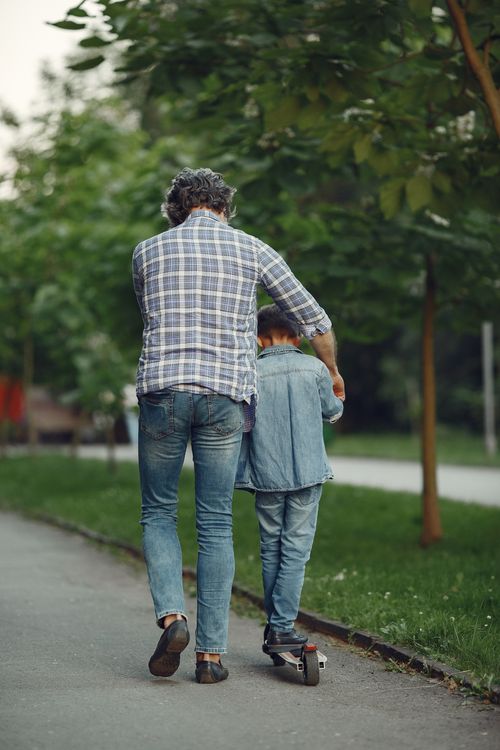 Free A man helps his son ride a skateboard along a tree-lined path. Stock Photo