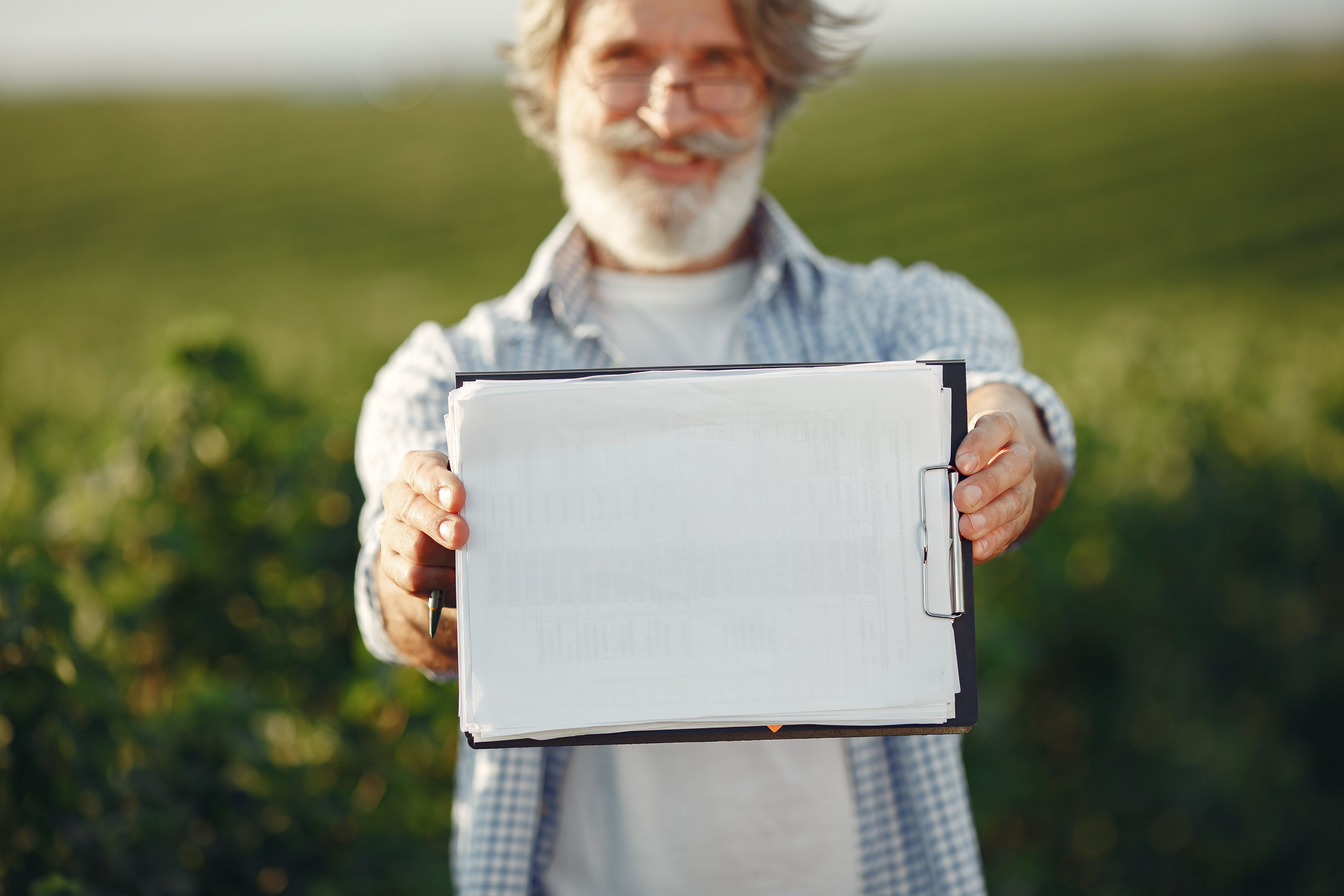 Bearded senior man with glasses holding a clipboard in a green field on a sunny day.